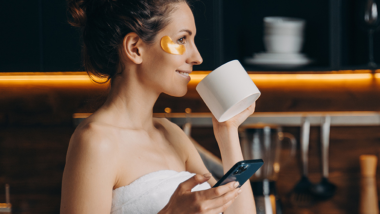 Frau mit Augenpads genießt Kollagen-Boost-Kaffee, während sie das Smartphone benutzt, Morgenroutine für Hautpflege und Entspannung.