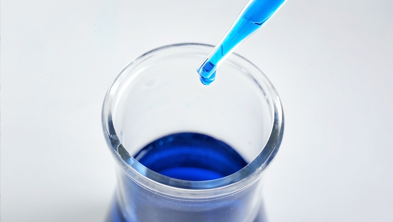 Ein Tropfen einer blauen Lösung wird in ein Becherglas pipettiert. Bildunterschrift: Der präzise Moment, wenn ein Tropfen blauer Farbstoff in ein klares Lösungsmittel fällt.